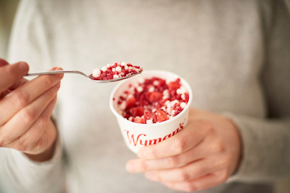 A person holding a cup labeled “Wyman’s” filled with a red and white frozen berry mixture, and lifting a spoonful of it.