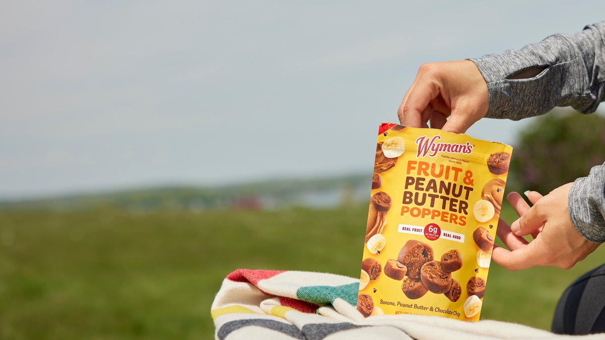 A hand picks up a piece of Wymans Fruit & Peanut Butter Poppers from a small plate next to a standing package on a kitchen counter.