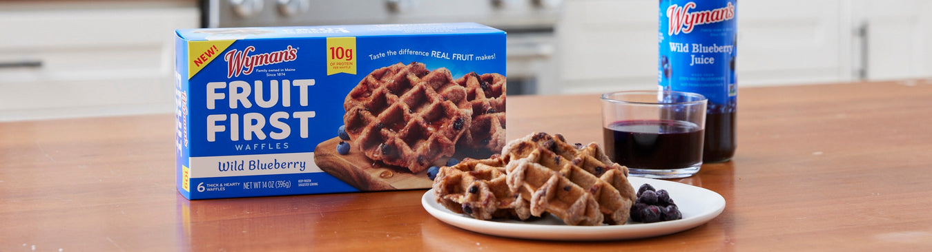 Plate of waffles with blueberries, a glass of orange juice, and a syrup dispenser on a wooden table. A green plant is blurred in the background.