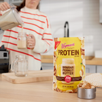 A pouch of Shop Wymans Protein Blend - Banana Peanut Butter rests on a kitchen counter as someone pours a protein smoothie into a jar in the background.
