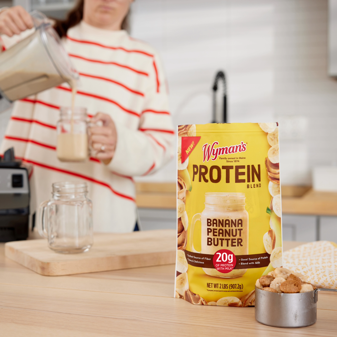 A pouch of Shop Wymans Protein Blend - Banana Peanut Butter rests on a kitchen counter as someone pours a protein smoothie into a jar in the background.