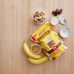 A bag of Shop Wyman’s Protein Blend - Banana Peanut Butter sits among bananas, peanut butter, banana slices, dates, milk, and flour—ideal for making a delicious protein smoothie with flash frozen fruit.