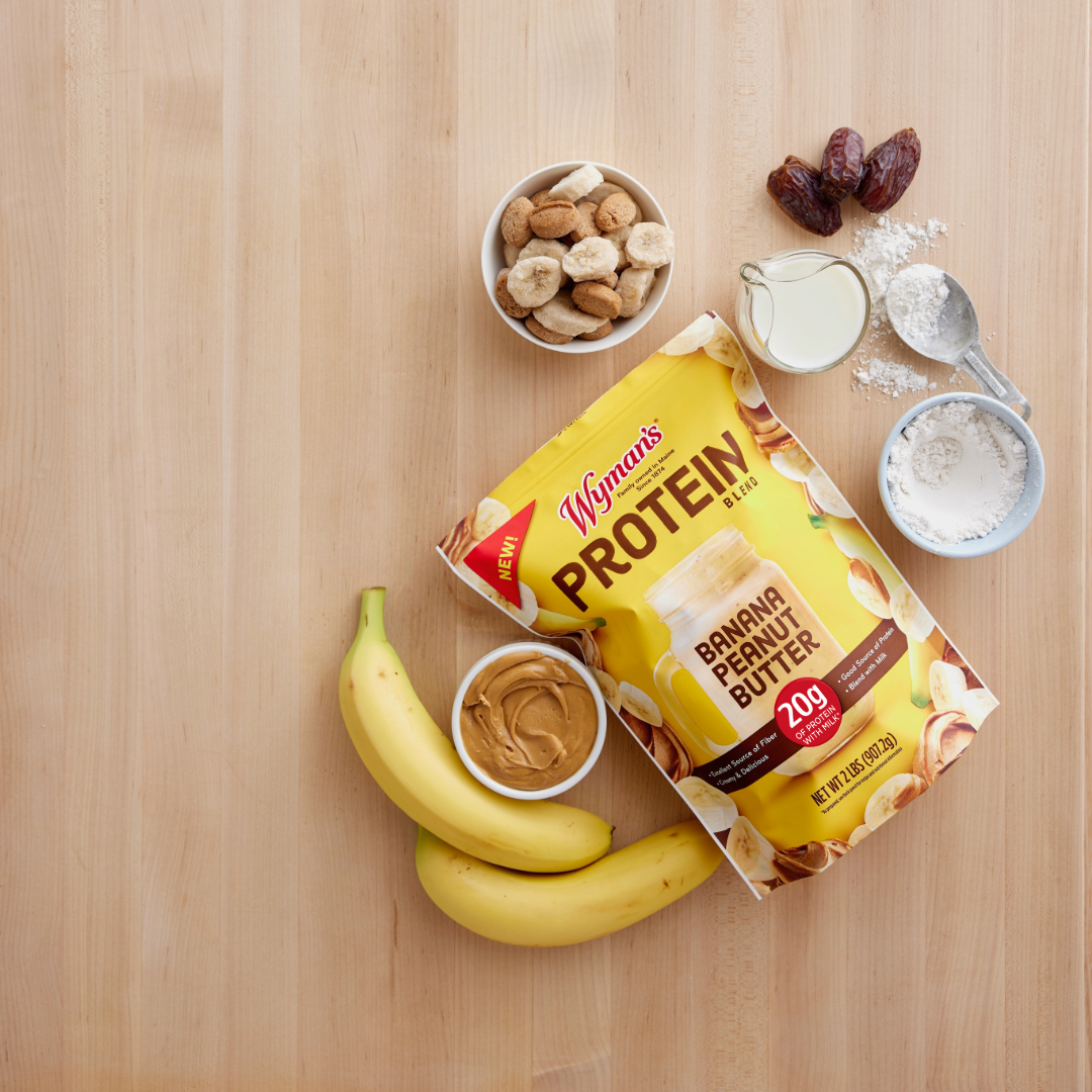 A bag of Shop Wyman’s Protein Blend - Banana Peanut Butter sits among bananas, peanut butter, banana slices, dates, milk, and flour—ideal for making a delicious protein smoothie with flash frozen fruit.