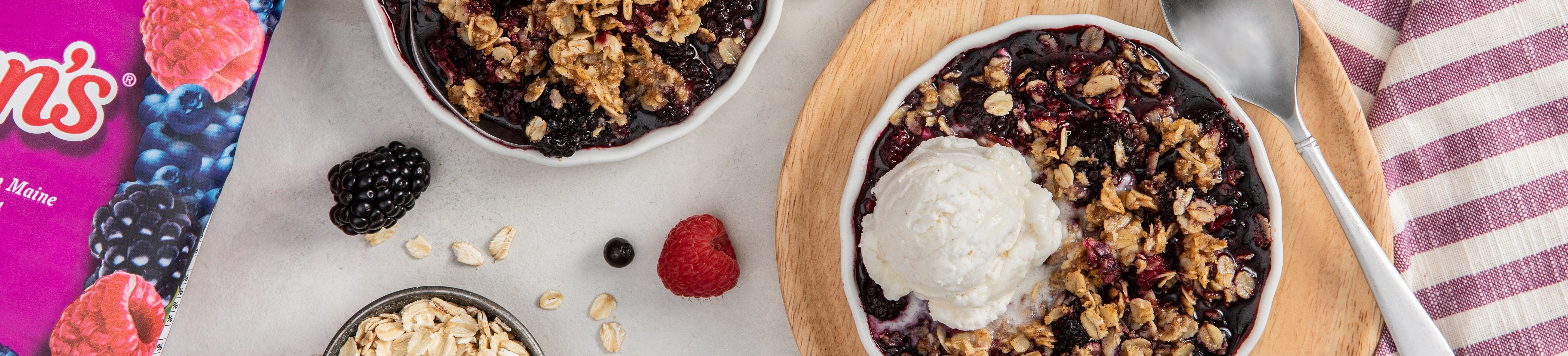 Two bowls of berry crumble, one topped with a scoop of vanilla ice cream, sit on a table with a spoon, fork, oats, loose berries, and a striped napkin beside a colorful berry package.