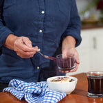 A person in a navy shirt sprinkles Wild Blueberry Powder - 8 oz bag over a dessert, holding its glass container. A blue and white checkered cloth and a glass of dark liquid sit on the wooden table.