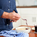 A person in a blue shirt sprinkles Wild Blueberry Powder - 8 oz bag onto oatmeal on a wooden table, next to a blue checkered cloth, a glass of dark liquid, and a small plate.