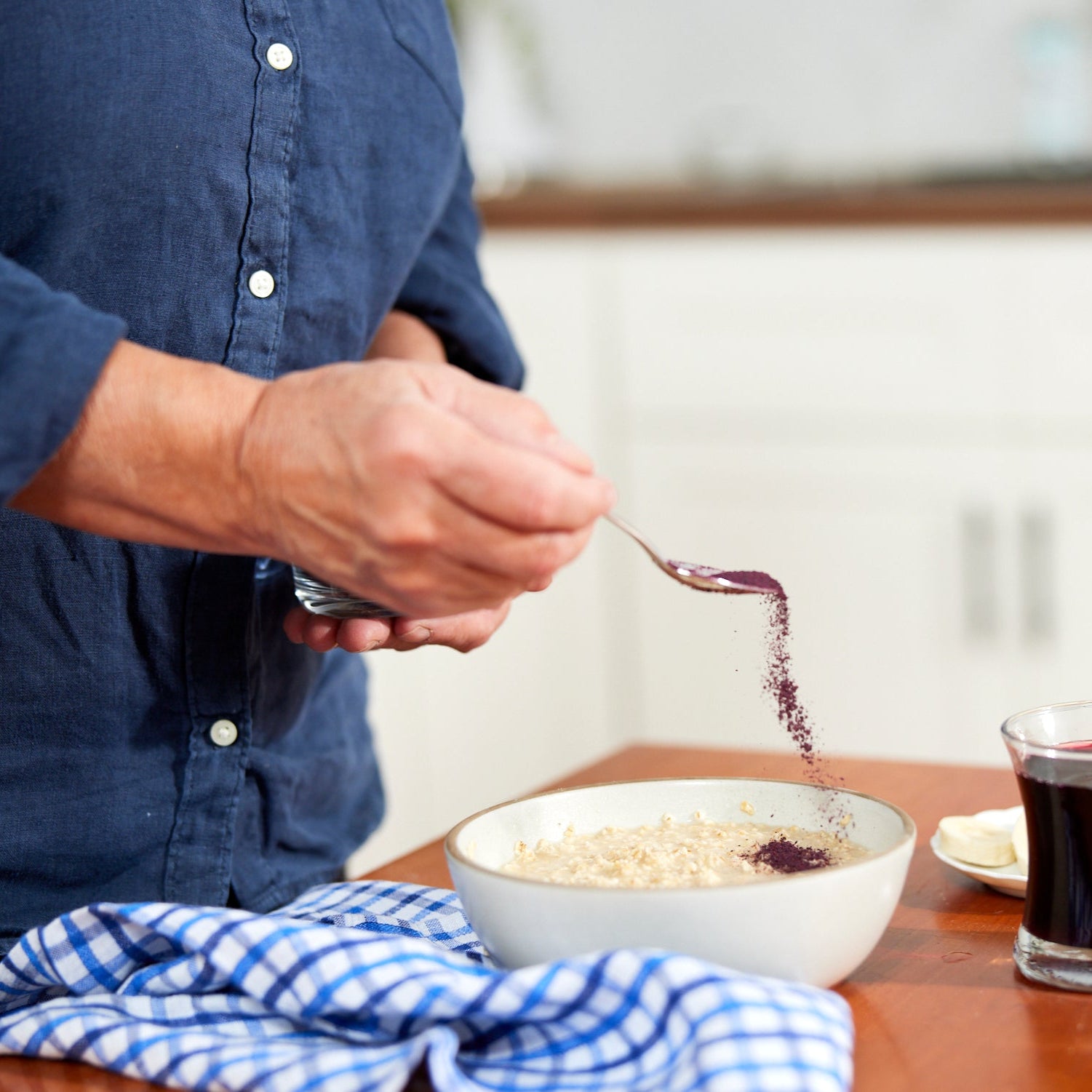 A person in a blue shirt sprinkles Wild Blueberry Powder - 8 oz bag onto oatmeal on a wooden table, next to a blue checkered cloth, a glass of dark liquid, and a small plate.