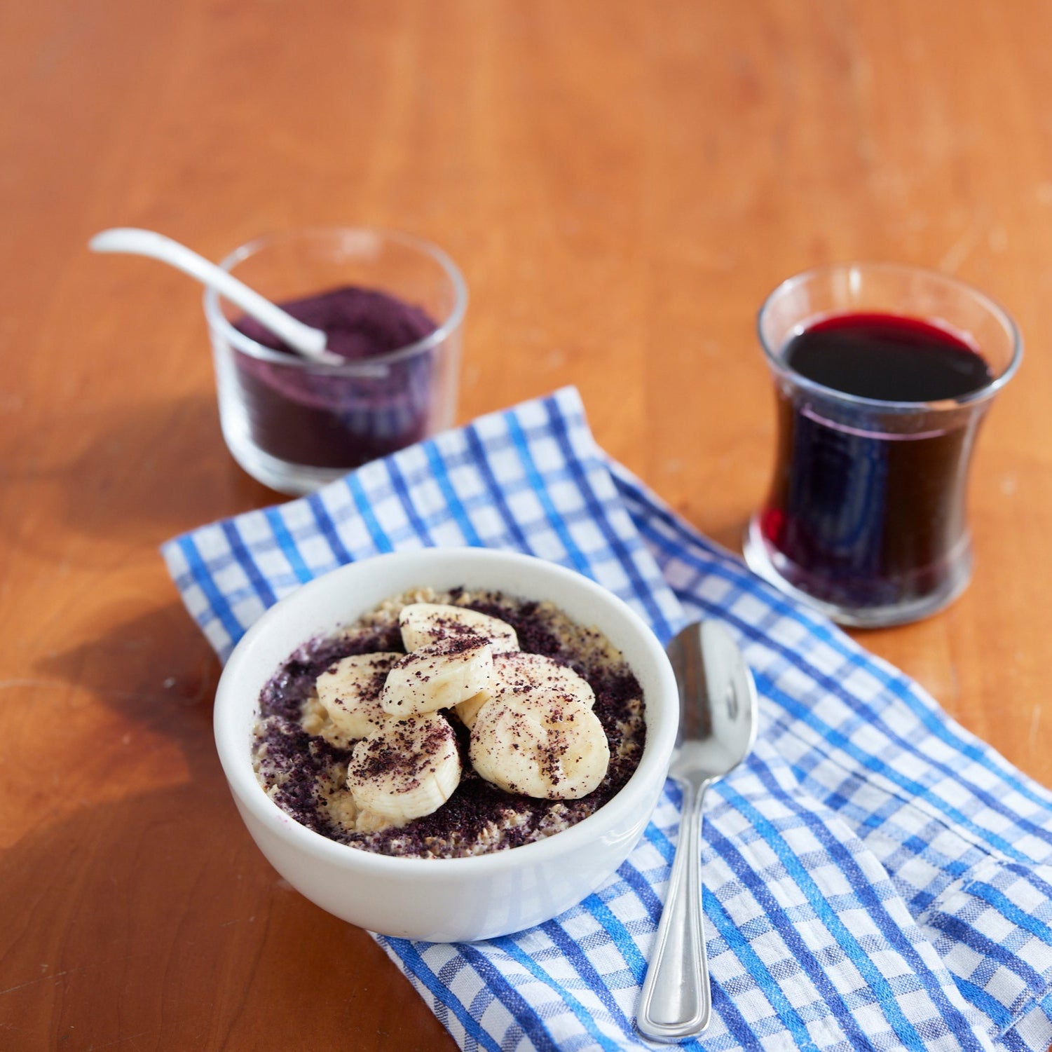 A white bowl of oatmeal topped with banana slices and Wild Blueberry Powder - 8 oz bag sits on a blue checkered cloth beside a spoon, with a glass of red juice and a small jar of fruit powder on a wooden table.