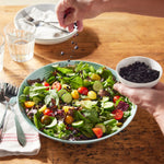 A hand sprinkles Whole Dried Wild Blueberries - 4.5 oz bag from a white bowl onto a fresh salad with cherry tomatoes, cucumbers, and greens in a blue bowl on a wooden table set with utensils and glasses.