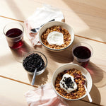 Two bowls of granola with yogurt and Whole Dried Wild Blueberries - 4.5 oz bag sit on a light wooden table, next to Wild Blueberry Juice, dried berries, and cloth napkins as sunlight casts soft shadows across the scene.