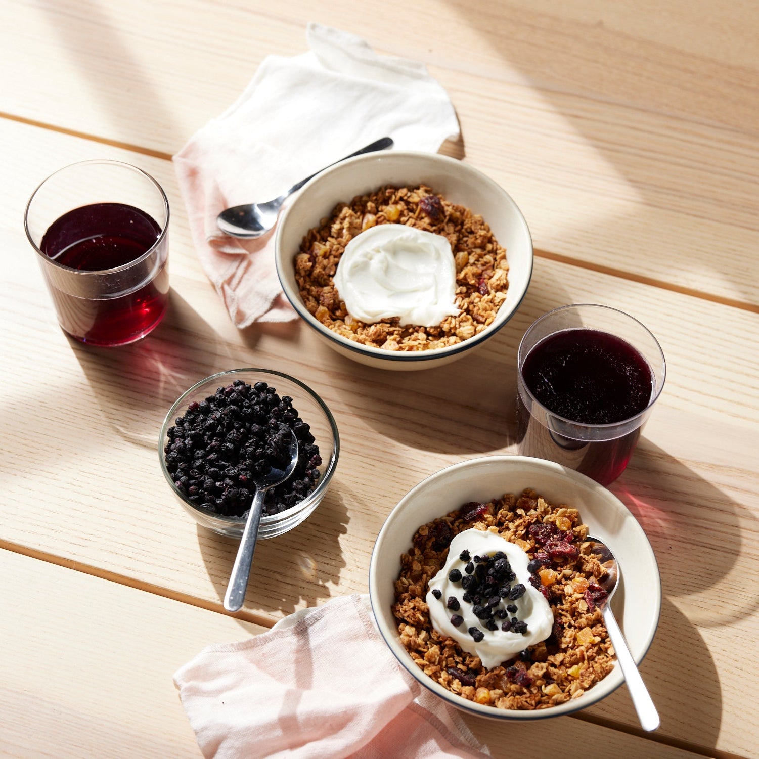 Two bowls of granola with yogurt and Whole Dried Wild Blueberries - 4.5 oz bag sit on a light wooden table, next to Wild Blueberry Juice, dried berries, and cloth napkins as sunlight casts soft shadows across the scene.