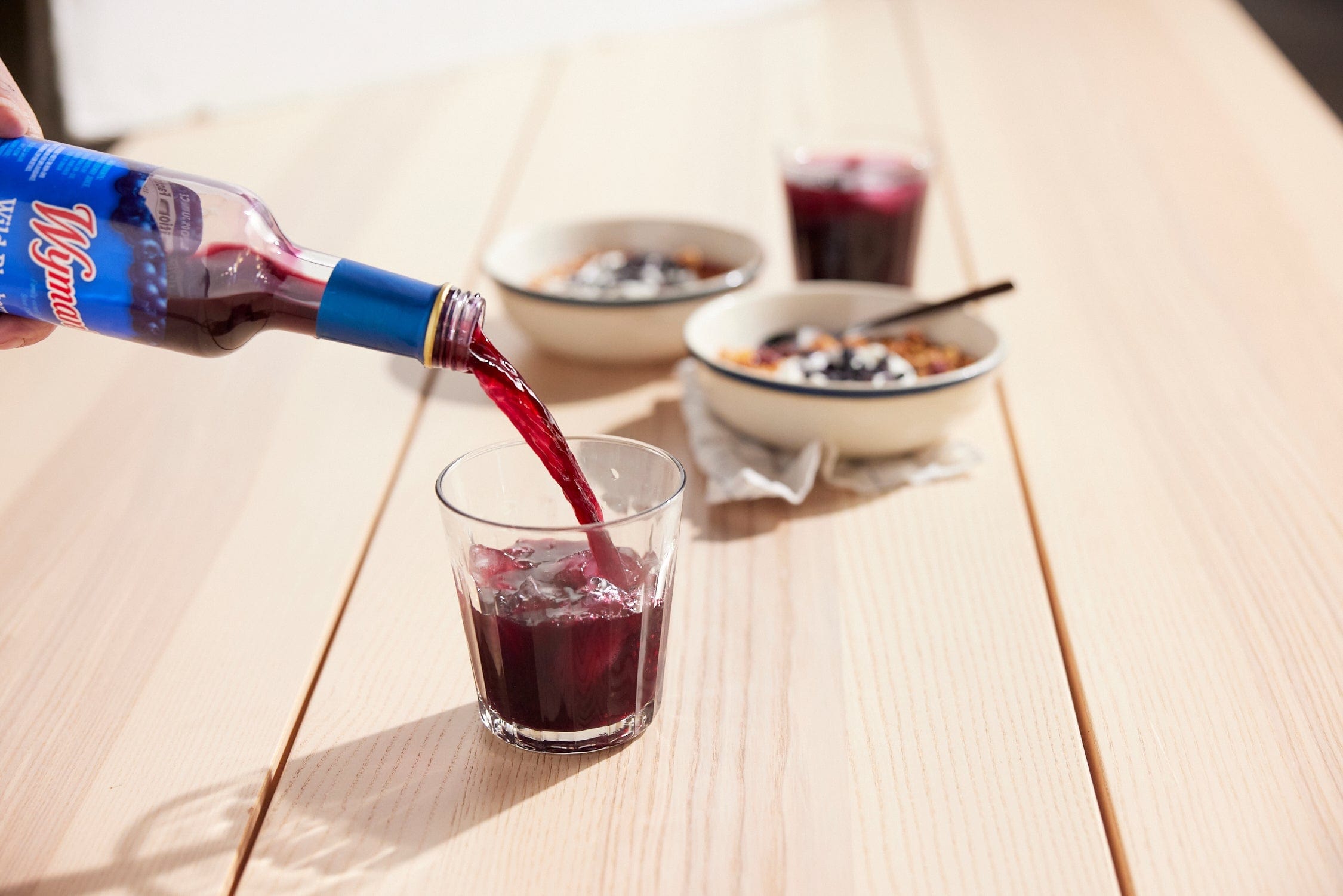 A hand pours a dark red beverage from a bottle into a glass filled with ice on a light wooden table, with two bowls of food and another glass in the background.