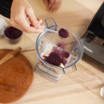 A hand holds a spoonful of Wild Blueberry Powder - 8 oz bag above a blender with ingredients, with a cutting board and blender base resting on a light wooden surface nearby.