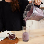 A person in a black sweater pours a thick purple smoothie—made with Wild Blueberry Powder - 8 oz bag—from a blender into a tall glass on a wooden table, with a small bowl of smoothie and a spoon beside it.