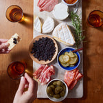 A wooden table holds a charcuterie board with assorted cheeses, sliced meats, olives, pickles, crackers, and Whole Dried Wild Blueberries - 4.5 oz bag. Two hands prepare a cracker snack with cheese and wild blueberry. Three amber drinks sit nearby.