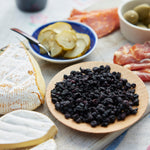 A wooden plate of Whole Dried Wild Blueberries - 4.5 oz bag is surrounded by soft cheese slices, pickles in a blue-rimmed bowl, green olives in a white dish, and pieces of cured meats on a serving board.