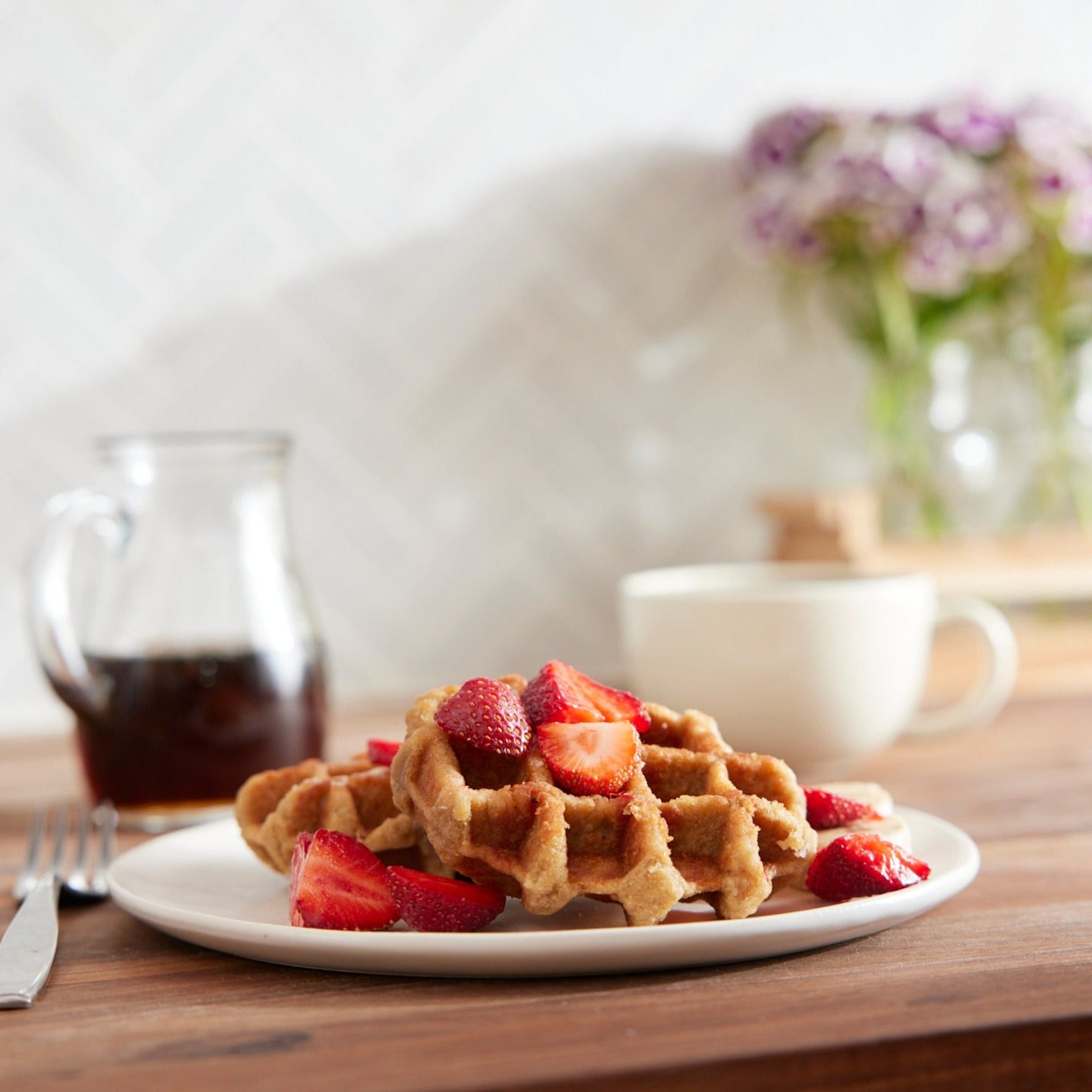 A plate of Shop Wyman's Fruit First Waffles - Homestyle topped with sliced strawberries, next to a fork, a jar of syrup, a cup of coffee, and a vase of flowers on a wooden table. Each waffle boasts 10g of protein.